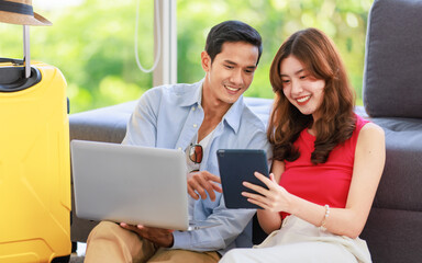 Asian young lovely boyfriend and girlfriend traveler couple sitting smiling on carpet in living room with trolley luggage while using laptop computer and tablet booking vacation summer trip online