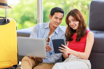 Asian young lovely boyfriend and girlfriend traveler couple sitting smiling on carpet in living room with trolley luggage while using laptop computer and tablet booking vacation summer trip online