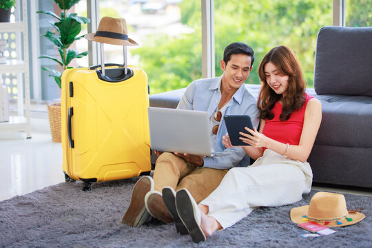Asian young lovely boyfriend and girlfriend traveler couple sitting smiling on carpet in living room with trolley luggage while using laptop computer and tablet booking vacation summer trip online