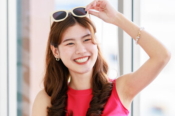 Asian young happy cheerful female traveler in casual summer outfit with sunglasses sitting smiling on cozy sofa in living room holding passport and boarding pass ticket with yellow trolley luggage