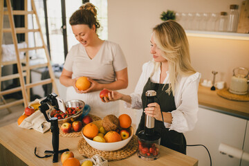 Two millennial woman bloggers recording video of cooking smoothie using smartphone on the kitchen.