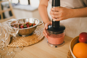 Millennial woman making strawberry smoothies using blender at the kitchen.