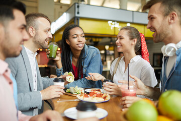 Group of cheerful young multi-ethnic students in casual clothing sitting at table on food court and having lunch together
