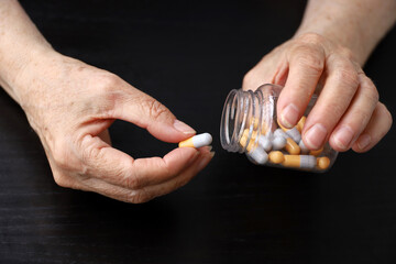 Pills in wrinkled hands of elderly woman on dark wooden table background. Medication in capsules,...