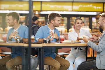 Cheerful emotional young people sitting at high table and talking while eating fast food in modern cafe
