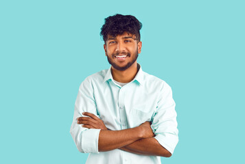 Portrait of happy ethnic student. Cheerful young dark skinned Indian, Arab, Gypsy or Bangladeshi man with curly black hair in white shirt standing with his arms folded, looking at camera and smiling - obrazy, fototapety, plakaty