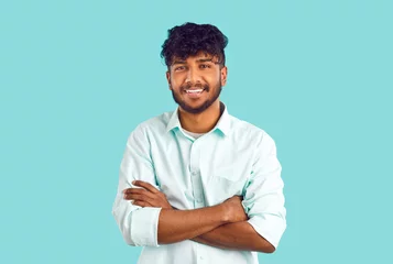Poster Portrait of happy ethnic student. Cheerful young dark skinned Indian, Arab, Gypsy or Bangladeshi man with curly black hair in white shirt standing with his arms folded, looking at camera and smiling © Studio Romantic