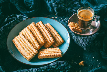 Traditional homemade shortbread cookies called “galletones manchegos” on a round blue plate next to a glass cup of coffee on a dark table. Sweet food for breakfast or snack.