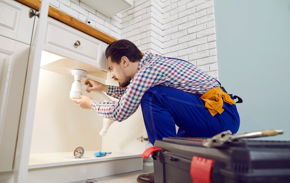 Plumber fixing problems in the kitchen. Man in an overall work uniform crouching on the floor beside the sink drain, trying to unclog the blockage, and checking, repairing or replacing some pipe parts
