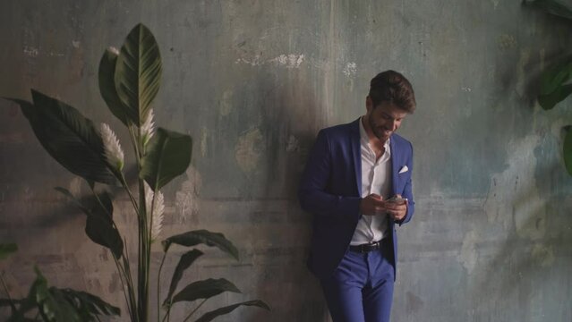 young elegant man in blue suit using mobile phone - businessman leaning against wall of ancient villa writing a message with his mobile phone - smiling attitude