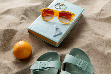 leisure and summer holidays concept - slippers, orange, earbuds, book and sunglasses on beach sand