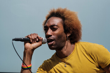 Non-binary young person is singing with a microphone in the street. He has a blue background.