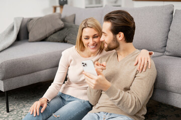 Caucasian couple with smartphone on the floor spending leisure time in social networks, scrolling feed, watching videos. Bearded guy is showing something to his girlfriend