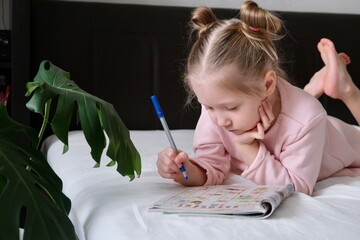 Portrait of a schoolgirl solves children crosswords on the bed. Logic games at home. Children...