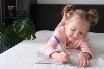 Portrait of a schoolgirl solves children crosswords on the bed. Logic games at home. Children...