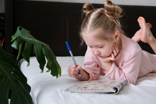Portrait of a schoolgirl solves children crosswords on the bed. Logic games at home. Children solves crossword