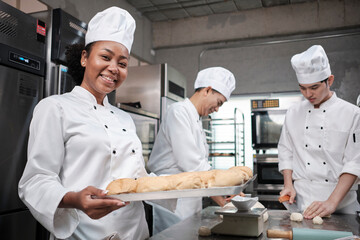 Portrait of African American female chef in white cooking uniform looking at camera with cheerful smile and proud with tray of bread in kitchen, pastry foods professional and fresh bakery occupation.