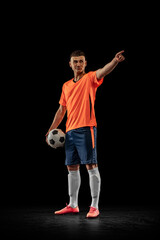 Young male soccer player in orange-blue football kit posing with ball isolated on dark background. Concept of sport, goals, competition, male hobby, occupations