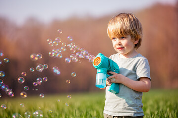 Little blond boy play soap bubbles gun stand in the field
