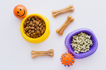 top view of bone shaped pet cookies near dry pet food in bowls and rubber toys on white.