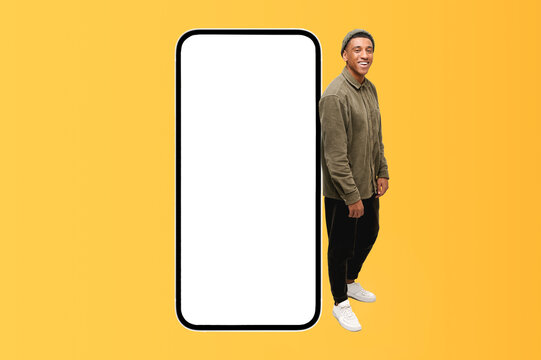 Best deal promotion. Smiling African-american teenage guy leaning on huge smartphone with empty blank screen, looking at the camera and smiles, hipster man advertising new app or website, mockup