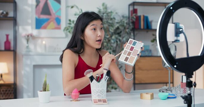 Experienced makeup artist of asian descent applies eye shadow on the eyelids, recording a tutorial on smoky eyes, in front of her on the table mirror, cosmetics and beauty accessories.