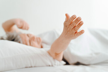 Fototapeta na wymiar Awakened senior caucasian woman stretches after sleep, pulls her hand to side while lying in bed in bedroom. Selective focus on female hand