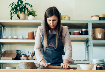Small Business: Serious woman potter using rolling pin for clay mass and making ceramics object on a desk in her studio