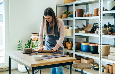 Small Business: Smiling woman potter using rolling pin for clay mass and making ceramics object on a desk in her studio