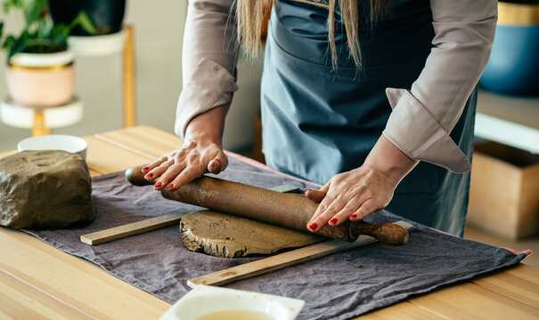 Close Up Photo Of Woman Hands Molding Clay At Her Workshop:  
Unrecognizable woman potter using rolling pin for clay mass and making ceramics object on a desk in her studio