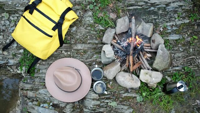 Top view of unrecognizable hiker young male prepare brunches for bonfire near river. Two mugs coffee, yellow backpack, hat and traveler man making campfire after hiking. Travel, lifestyle concept
