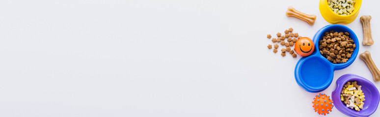 top view of bone shaped treats near bowls with dry pet food and rubber toys isolated on white, banner.