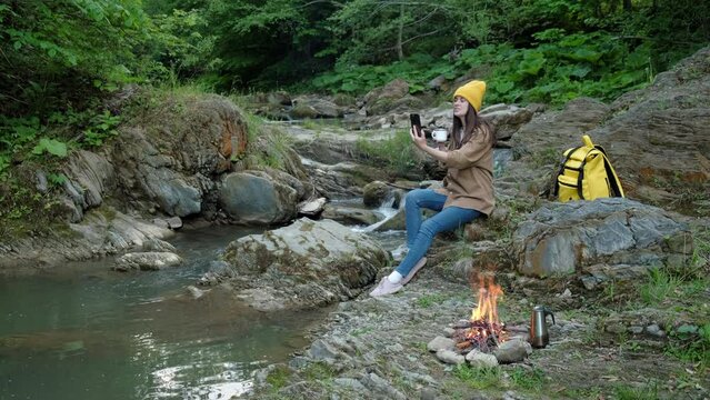 Adventurous millennial or hipster teenager, young pretty woman in knit beanie, sits on stones near mountain river uses smartphone to talk to friends. Hiker girl recording stories for social media