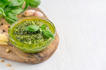 Italian traditional herbal pesto sauce made of blended parmesan cheese, basil leaves, pine nuts, garlic and olive oil served in glass bowl on cutting board on white wooden background with copy sapce