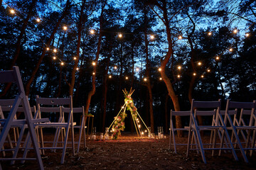 Festive string lights illumination on boho tipi arch decor on outdoor wedding ceremony venue in pine forest at night. Vintage string lights bulb garlands shining above chairs at summer rural wedding. © artiemedvedev