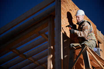 Carpenter hammering nail into OSB panel on the wall of future cottage at night. Man worker building...