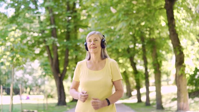 Senior active gray-haired woman jogging in urban city summer park outdoor. Mature old fitness female running exercise with headphones enjoys and listens to music. Elderly sport runner Workout cardio