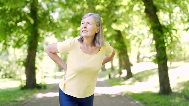 senior woman back pain in the park outdoors while jogging. Worried upset mature female feel hurt sudden ache. Upset eldery person suffering backache, unhappy lady lumbar hurt in the spine or lower