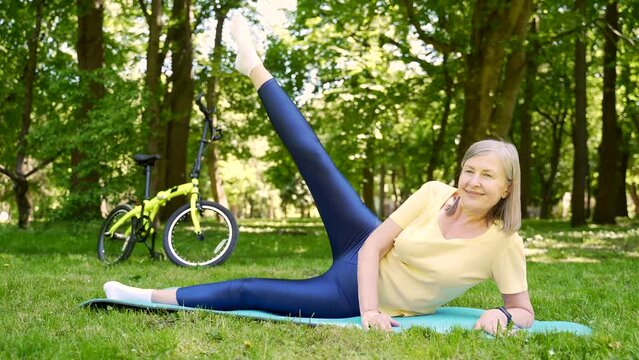 Active Gray Hair senior fitness woman doing stretching exercise on yoga fit mat outdoors in the city park on the grass. Mature modern female Elderly and healthy lifestyle Activity sports concept