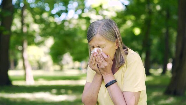 Senior elderly woman in the park with allergies has a runny nose and sneezes near trees blowing in handkerchief. Sick old female with tissue allergy sneezes outdoors cough In the park in nature