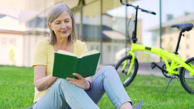 Senior woman reading a book on a meadow sitting on the grass lawn in an urban city park. Outdoors. Modern happy mature female spends leisure time in nature. Rest calm on fresh air. Carefree lifestyle
