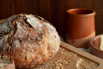 Homemade bread with cereals of round shape is located on a cutting board against the background of ceramic dishes. Rustic style.