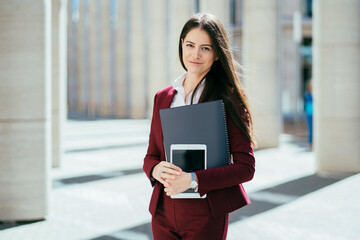 Attractive brunette elegant business woman in white shirt and burgundy suit, looking confident, standing over blurry city background. Business and education concept.