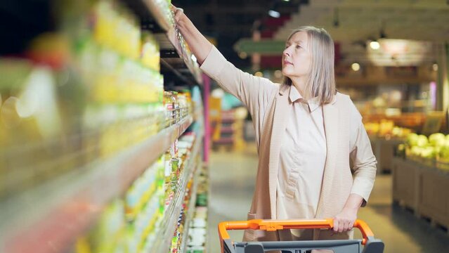 Senior retired woman is looking for and choosing product in a grocery store. Mature female supermarket shopper inspects and pick shelves. has difficulty choosing the range search goods in food market