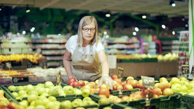 A senior female greengrocer worker in a supermarket or grocery store or a small business owner picks apples near the fruit department in the market. Mature female employee working behind the counter