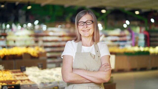 Portrait senior mature woman worker in a Vegetable section supermarket standing smiling with arms crossed. Friendly pleasant female looking at camera in a fruit shop market. Employee greengrocer
