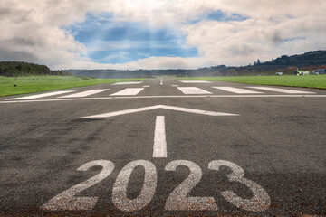White arrow on a small asphalt air plane runway and sign 2023. Cloudy sky with small blue opening in the middle. Sun rays and flare. Small airport on the right. Selective focus