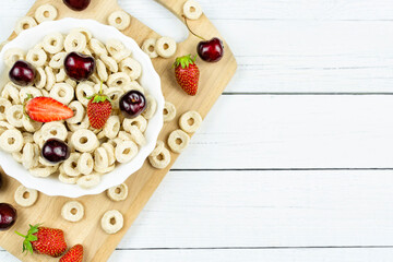 A bowl with oatmeal (corn) rings with strawberries and cherries on a wooden board on a white background. A place for the text, a recipe. Dry healthy breakfast, add milk.