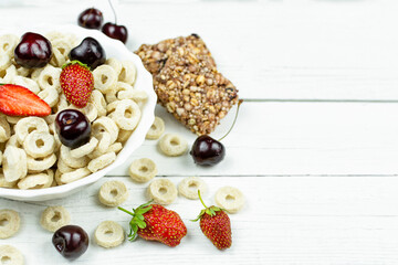 A bowl with oatmeal (corn, whole grain) rings with strawberries and cherries on a white wooden background. Space for the text. Dry healthy breakfast, add milk.