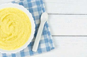 Corn porridge for a baby made of ground cereals in a white bowl on a blue cloth napkin on a white wooden background. Space for the text. The first complementary food of a child, baby nutrition.